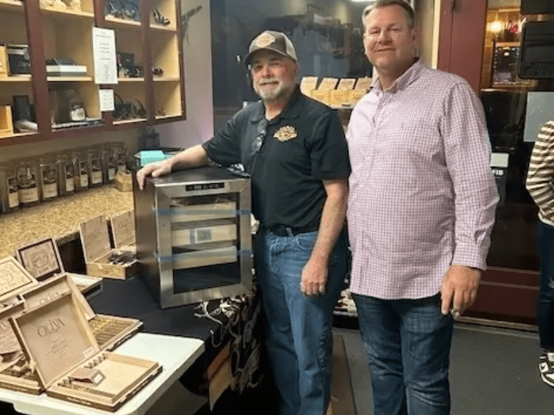 Two men stand beside a table, engaging in conversation next to an open box of cigars.