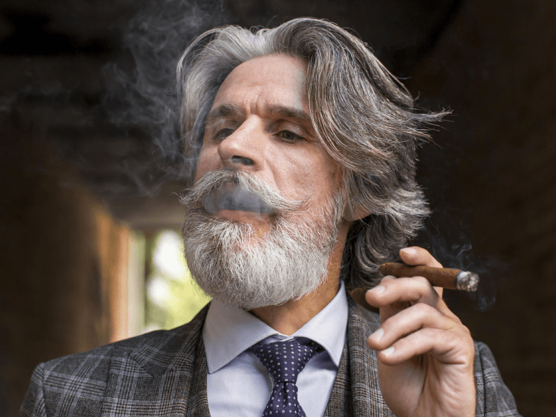 A bearded man in a suit confidently smokes a cigar, exuding sophistication and style in a refined setting.